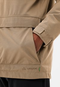 Tan waterdichte jas met hoge kraag, zijzak en klittenbandsluiting bij de manchetten. Kenmerkt de merknaam "Vaude" geborduurd op de zoom.