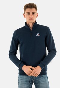 Navy blue zip-neck sweatshirt featuring a logo on the chest, long sleeves, and a fitted design. Made from a soft, smooth fabric.