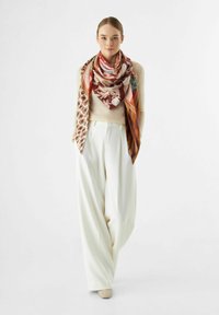 Écharpe en soie avec des motifs floraux et géométriques dans des tons chauds, associée à un pull léger et un pantalon blanc ample. Les chaussures s'harmonisent avec l'ensemble.
