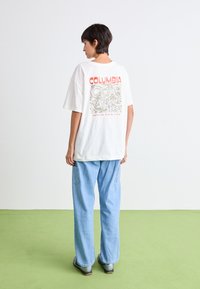 Bílý tričko s červenou grafikou "Columbia" a designem hory na zádech, doplněné světle modrými širokými džínami a šedými botami.