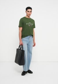 Muž v zeleném tričku Tommy Hilfiger, světlých modrých džínách, černých botách, držící velkou černou koženou kabelku, stojící na bílém pozadí.