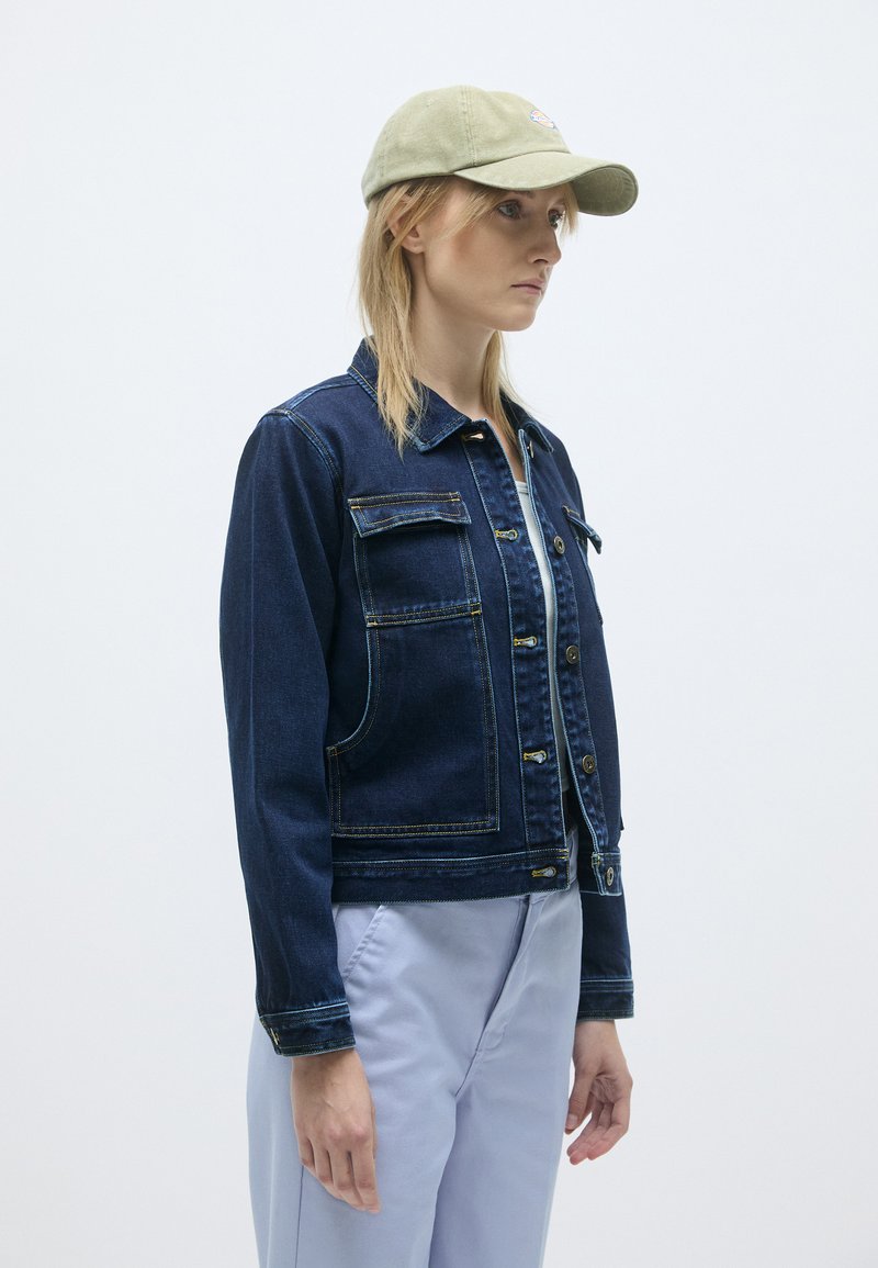 Donkerblauwe spijkerjack met gouden stiksels, voorzien van twee voorkruiszakken en knoopsluitingen. Vergezeld van een lichtgroene pet.