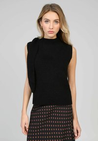 Pull noir sans manches avec un col montant et un détail drapé sur une épaule, texture tricotée, associé à une jupe marron à pois.