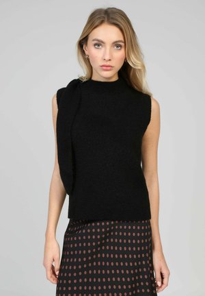 Pull noir sans manches avec un col montant et un détail drapé sur une épaule, texture tricotée, associé à une jupe marron à pois.