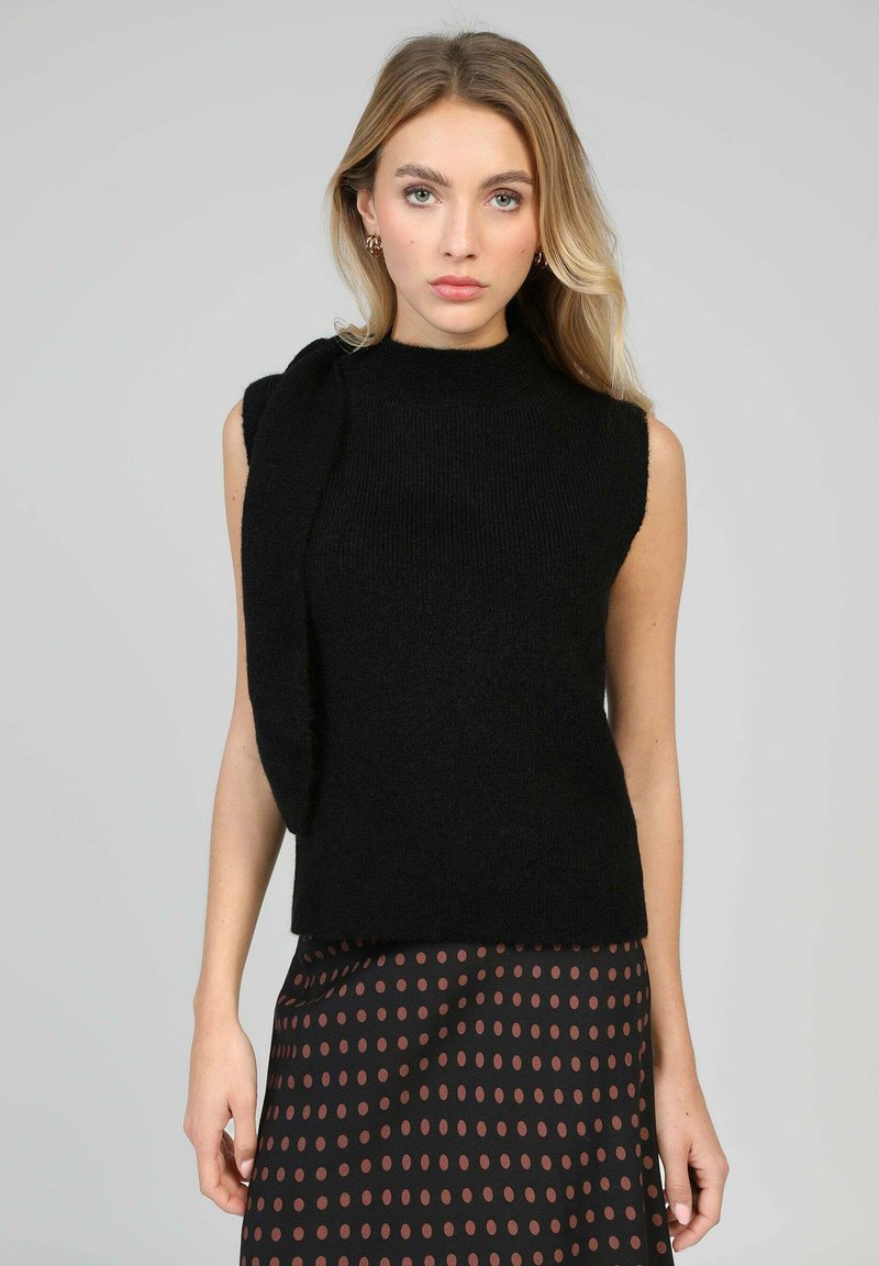 Pull noir sans manches avec un col montant et un détail drapé sur une épaule, texture tricotée, associé à une jupe marron à pois.