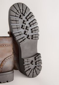 Brauner Lederstiefel mit einer strukturierten Gummisohle, die ein robustes Profil für besseren Halt aufweist. Verziert mit Brogue-Details entlang der Seiten.