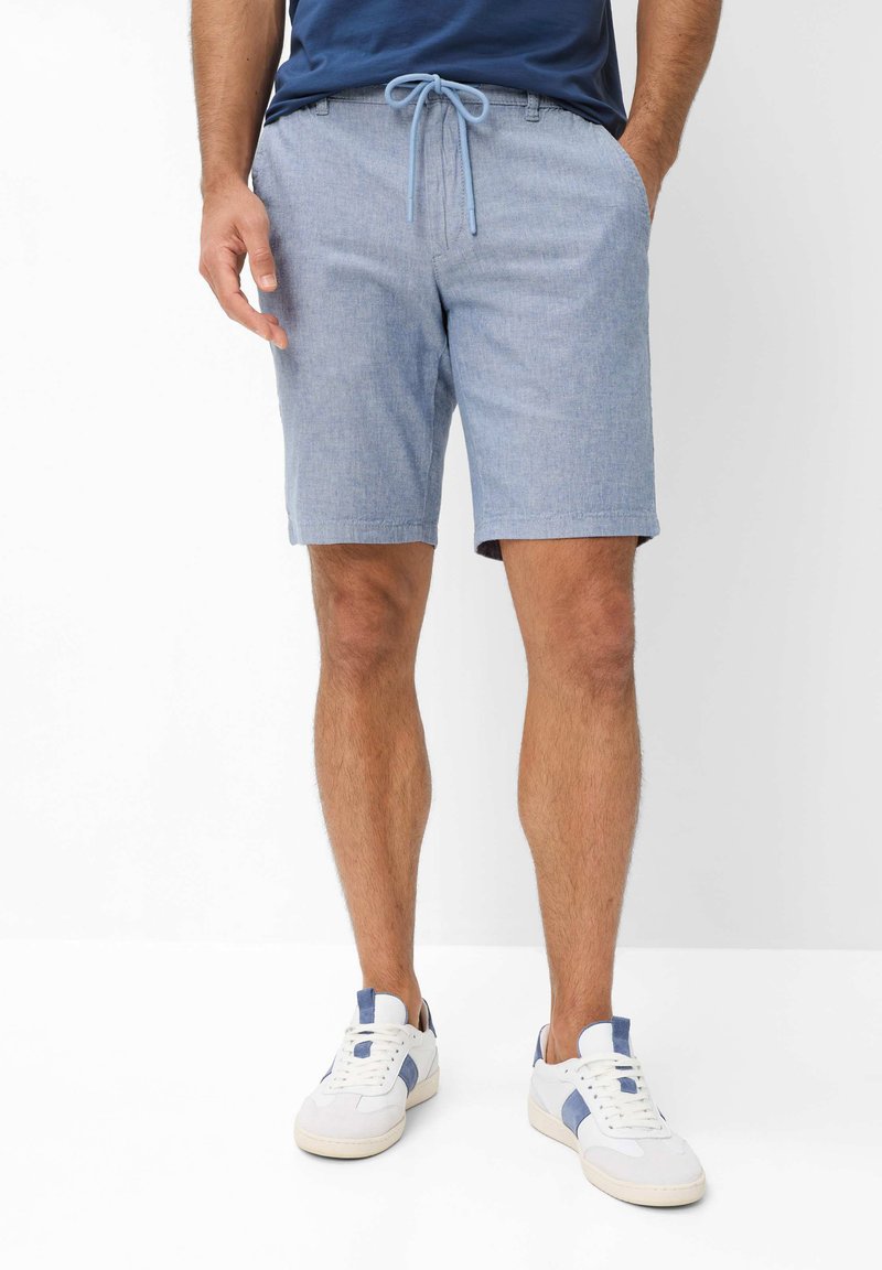 Man draagt lichtblauwe shorts met trekkoord, een blauw shirt en witte sneakers met blauwe accenten, staand tegen een effen witte achtergrond.