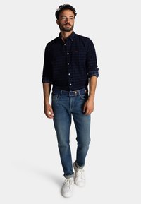 Marineblauwe geruite knop-overhemd met lange mouwen, gecombineerd met middelblauwe jeans en witte sneakers. Beschikt over een contrasterende riem. Eenvoudig ontwerp.