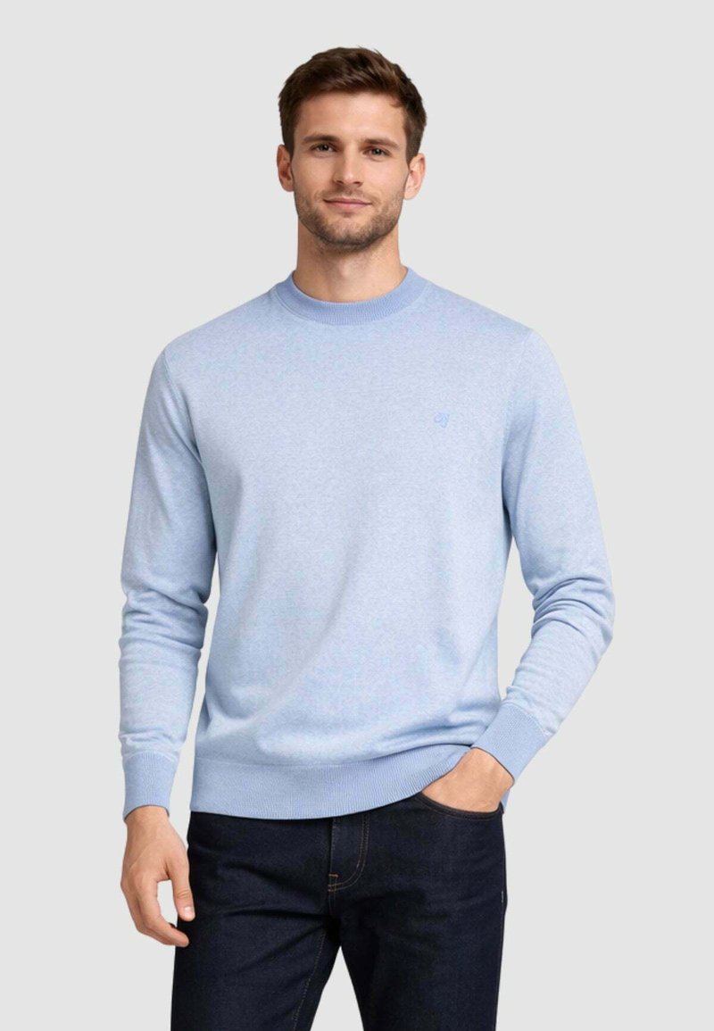 Hombre con un suéter azul claro de cuello redondo y vaqueros oscuros, de pie con una mano en el bolsillo frente a un fondo liso.