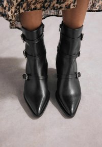 Botines de cuero negro con punta afilada, que presentan tres correas con hebillas de metal y una textura suave.