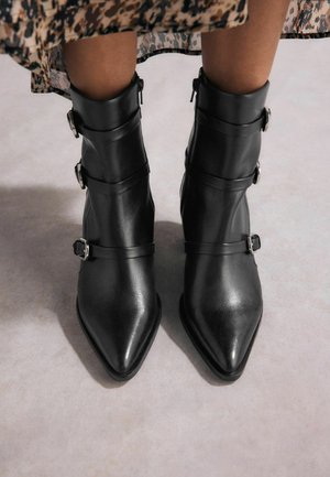 Botines de cuero negro con punta afilada, que presentan tres correas con hebillas de metal y una textura suave.