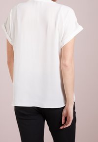 Kvinna som bär en kortärmad vit blus med uppvikta ärmar och svarta byxor, sedd bakifrån mot en neutral bakgrund.