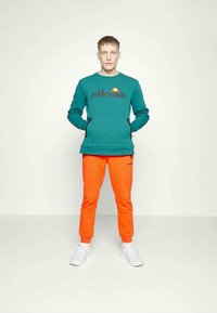 Felpa teal con logo, dettagli neri e zip laterali; abbinata a pantaloni arancioni a sigaretta; il modello indossa sneaker bianche.