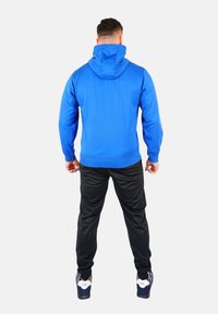 Sudadera azul con una gran capucha, puños y dobladillo acanalados, combinada con pantalones deportivos negros y zapatillas. Tejido suave, diseño ajustado.