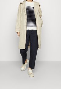 Parka lunga beige, maglione lavorato a maglia a righe navy, pantaloni navy e sneakers sportive color crema, evidenziando morbidezza e design a strati.