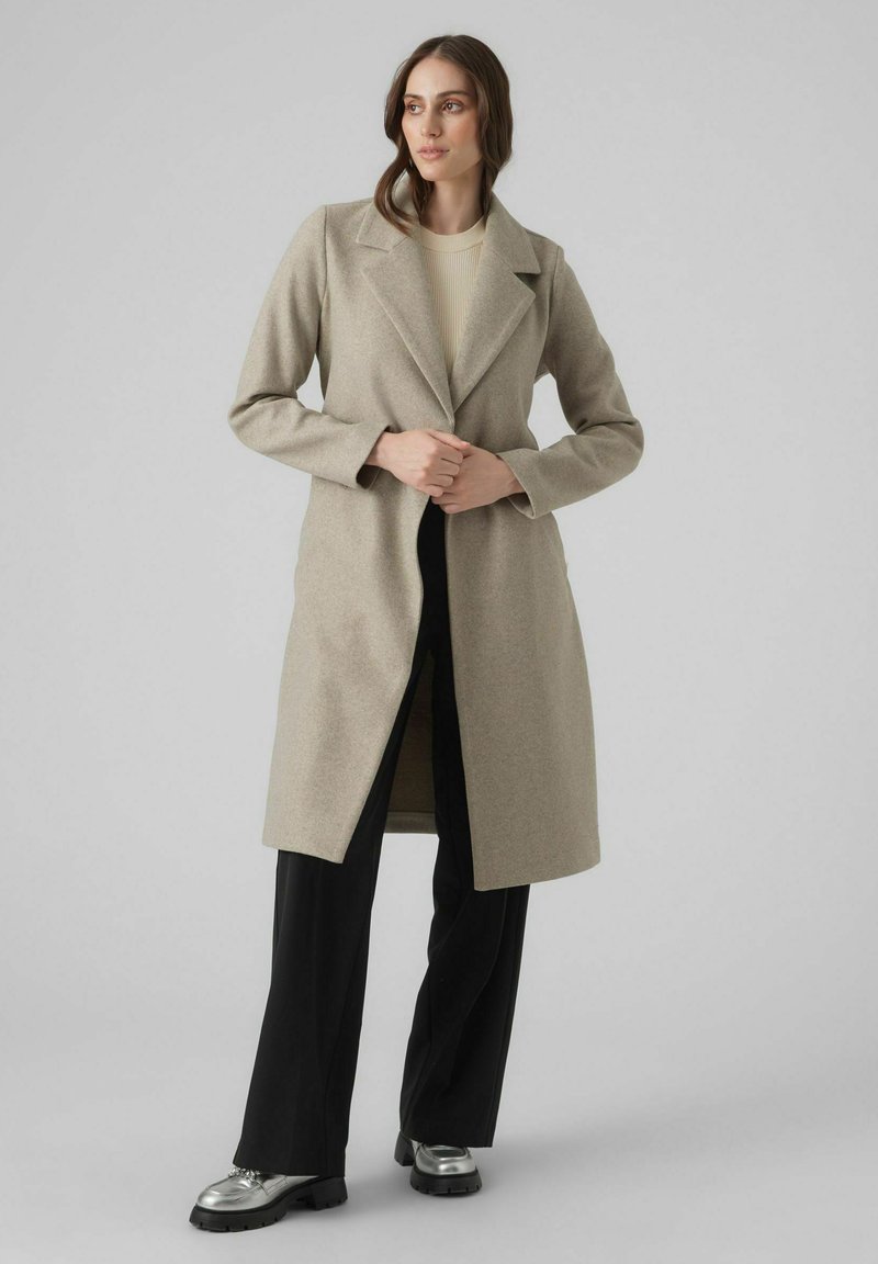 Cappotto lungo in misto lana beige con colletto a punta, apertura frontale e maniche lunghe, abbinato a pantaloni neri e scarpe metalliche.