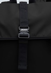 Mochila negra con un diseño estructurado, que cuenta con una correa texturizada y un cierre de plástico etiquetado como "JACKIT". Superficie de tela suave.