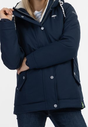 Femme portant une veste d'hiver bleu marine avec capuche, boutons devant, poches latérales et doublure en polaire, debout les bras croisés.