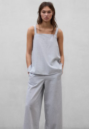 Mujer joven de pie con las manos en los bolsillos, vistiendo una blusa sin mangas y suelta y pantalones de pierna ancha con finas rayas verticales sobre un fondo gris liso.