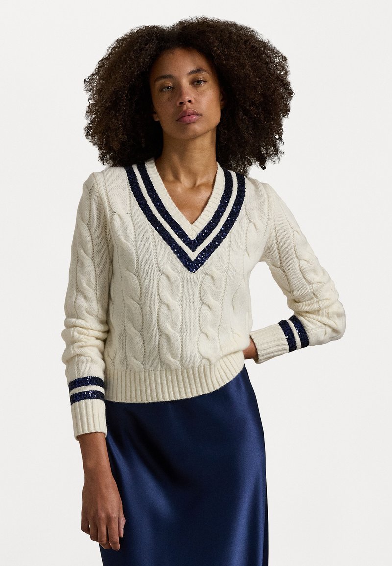 Pull en maille torsadée crème avec un col en V, orné de rayures navy et d'accents scintillants au col et aux poignets, associé à une jupe satinée navy.