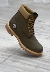 Boots de ancle maro, impermeabile, cu guler bej, sistem de închidere cu șireturi, talpă din cauciuc și patch cu logo pe lateral. Material texturat și cusături întărite.