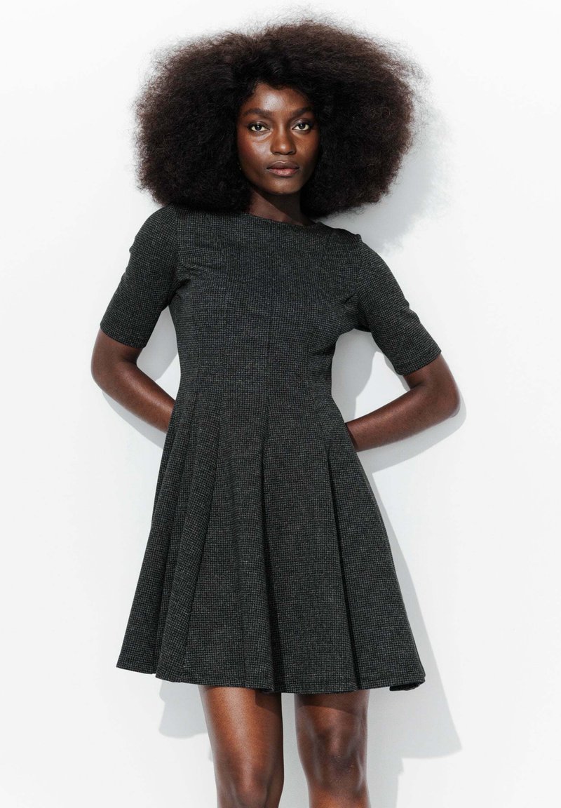 Robe noire structurée à manches courtes avec une jupe évasée, présentant un tissu texturé et des détails de plissage, sur fond blanc.