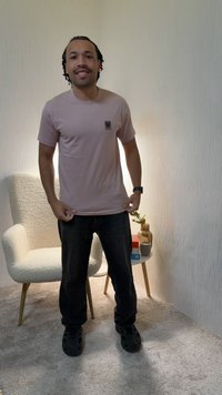 Homme souriant, portant un t-shirt rose clair et un pantalon noir, debout dans une pièce avec moquette équipée d'un fauteuil blanc et d'une petite table avec un vase et des livres.