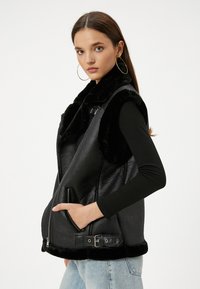 Schwarze Kunstlederweste mit einem weichen Fellkragen, Reißverschluss und Fronttaschen. Mit einem schwarzen Ledergürtel am Saum akzentuiert.