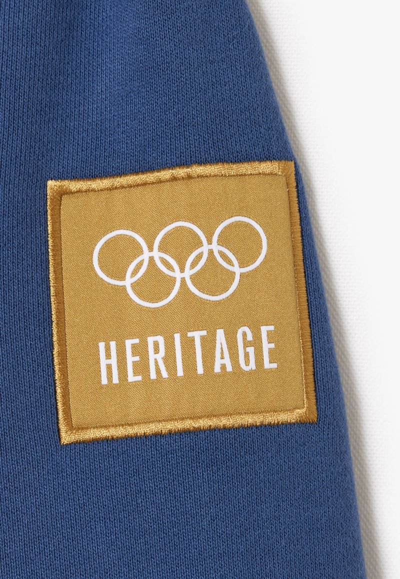 Blaues Gewebe mit einem goldenen rechteckigen Fleck. Der Fleck zeigt weißen Text "HERITAGE" und ein Design der olympischen Ringe in Weiß. Die Textur ist glatt und gestickt.