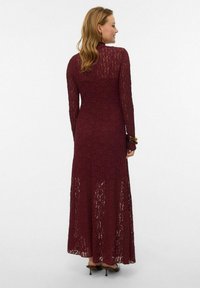 Viininpunainen pitsinen maksimekko, jossa on pitkät hihat, teksturoitu kukkakuvio ja vartalonmyötäinen siluetti. Mekossa on liehukehelma ja hillityt läpikuultavat yksityiskohdat.