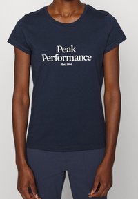 Námořnická modrá bavlněná tričko s krátkým rukávem, s bílým textem "Peak Performance" a "Est. 1986" ve středovém, pohodlném písmu.