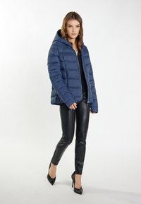 Giacca puffer blu navy con cappuccio, design trapuntato e chiusura con zip. Abbinata a pantaloni in pelle nera e tacchi alti, su uno sfondo bianco.