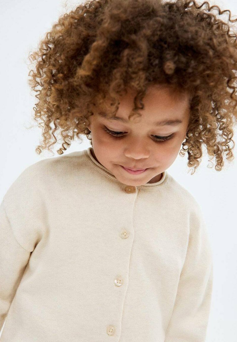 Cardigan bej din tricot cu guler rotund și închidere cu nasturi, având o textură moale și mâneci lungi, potrivit pentru copii.