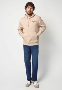 Sudadera beige con cordones, bolsillo frontal y logotipo impreso. Combinada con jeans azul oscuro y zapatillas blancas con detalles en gris.