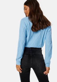 Světle modrý pletený svetr s diamantovými vzory, dlouhými rukávy a žebrovanými manžetami, spárovaný s černými džíny s vysokým pasem.