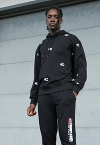 Felpa nera con stampa del logo bianca, cappuccio con cordino e polsini a costine, abbinata a pantaloni neri con testo grafico rosso sulla gamba.