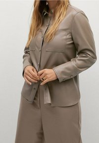 Světle hnědá koženka košile s knoflíkovou přední částí, dvěma náprsními kapsami a uvolněným střihem. Hladká textura a dobře střižené rukávy.