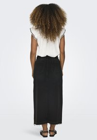 Zwarte denim maxi-rok met een rafelrand, hoge taille en twee achterzakken. Gecombineerd met een witte blouse met ruches aan de mouwen en zwarte bies.