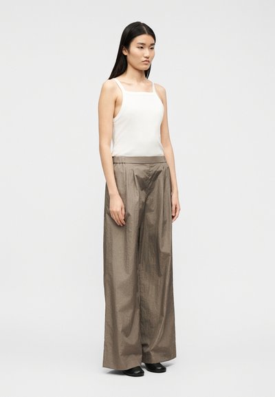 Nő áll, fehér ujjatlan felsőt, bő, bézs színű, széles szárú nadrágot és fekete cipőt visel, egyszínű világos háttér előtt.