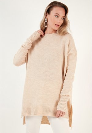 Beige long-sleeve sweater made of soft knit material, featuring a ribbed hem and side slits for a relaxed fit. Light texture throughout.