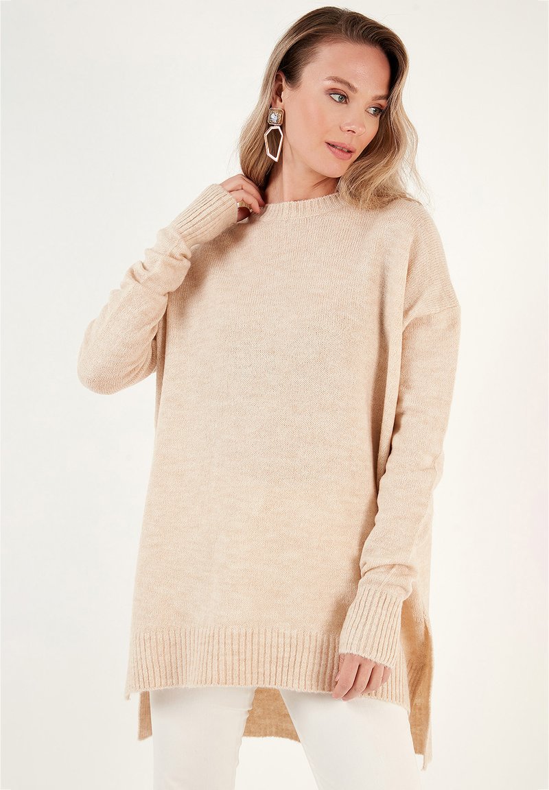 Pull à manches longues beige en tricot doux, présentant un ourlet côtelé et des fentes sur les côtés pour un ajustement décontracté. Texture légère partout.
