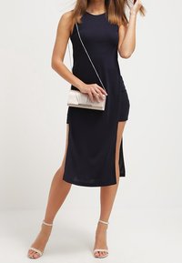 Kvinna i ärmlös svart klänning med hög slits, som håller en beige kuvertväska, iförd beige ankelremmsklackar mot en enkel vit bakgrund.