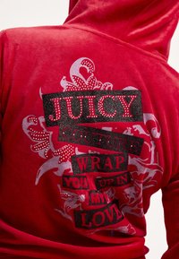 Piros velúr kapucnis pulóver, amely vastag fekete és csillogó szöveggel, valamint virágos díszítéssel rendelkezik. A mintán szerepelnek az „JUICY COUTURE” és a „WRAP YOU UP IN MY LOVE” feliratok.
