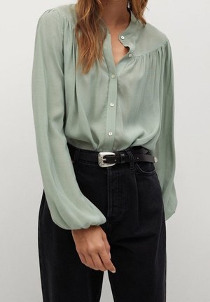 Femme portant un chemisier vert clair boutonné avec des manches bouffantes, rentré dans un jean noir taille haute avec une ceinture noire.