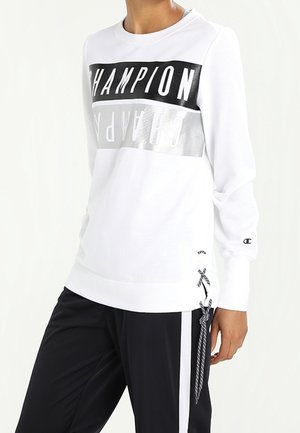 Osoba ubrana w białą bluzę Champion z czarnymi i srebrnymi pasami z logo oraz czarne spodnie z białymi bocznymi paskami i zawiązanym ściągaczem.