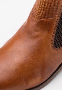 Hnědé kožené kotníkové boty s texturovaným vzorem na svršku a elastickými akcenty. Hladký povrch s kulatou špičkou a švy.
