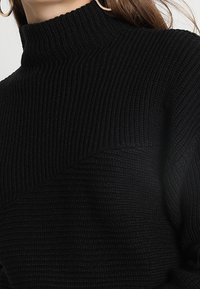 Czarny sweter z dzianiny z wysokim, ściąganym kołnierzem i fakturowanym wzorem. Materiał wydaje się miękki i ciepły, charakteryzuje się luźnym krojem.