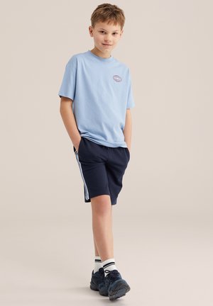 Băiat stând cu mâinile în buzunare, purtând un tricou bleu, pantaloni scurți bleumarin cu dungi laterale, șosete albe și adidași negri, pe un fundal bej.