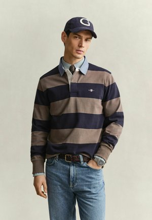 Junger Mann mit gestreiftem Rugby-Shirt, Jeans, einer blauen Kappe mit dem Buchstaben G und einer gemusterten Krawatte unter dem Kragen.