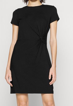 Žena v krátkých rukávech černých šatech s uzlem v pase, stojící před jednobarevným světlým pozadím.